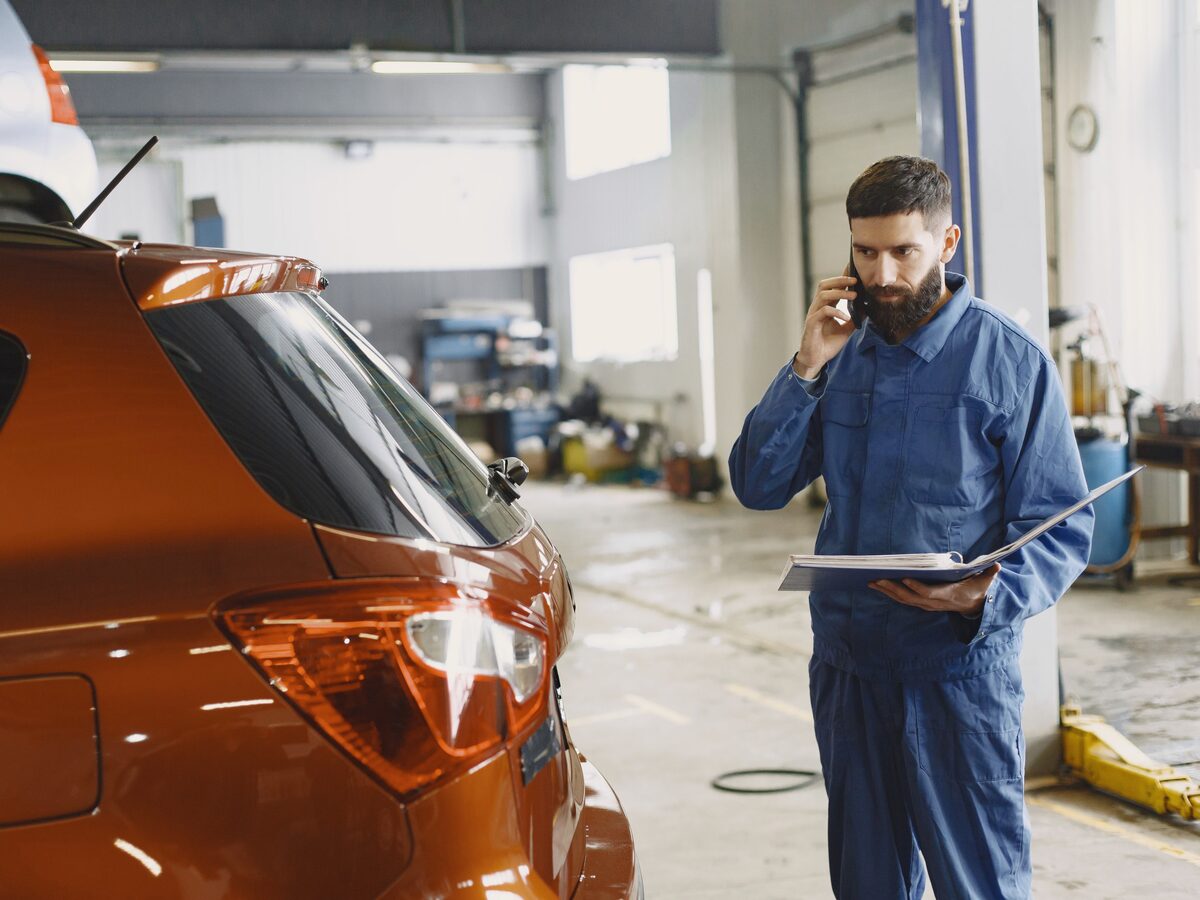 A mechanic on a phone call in his workshop, holding a job folder beside a customer car